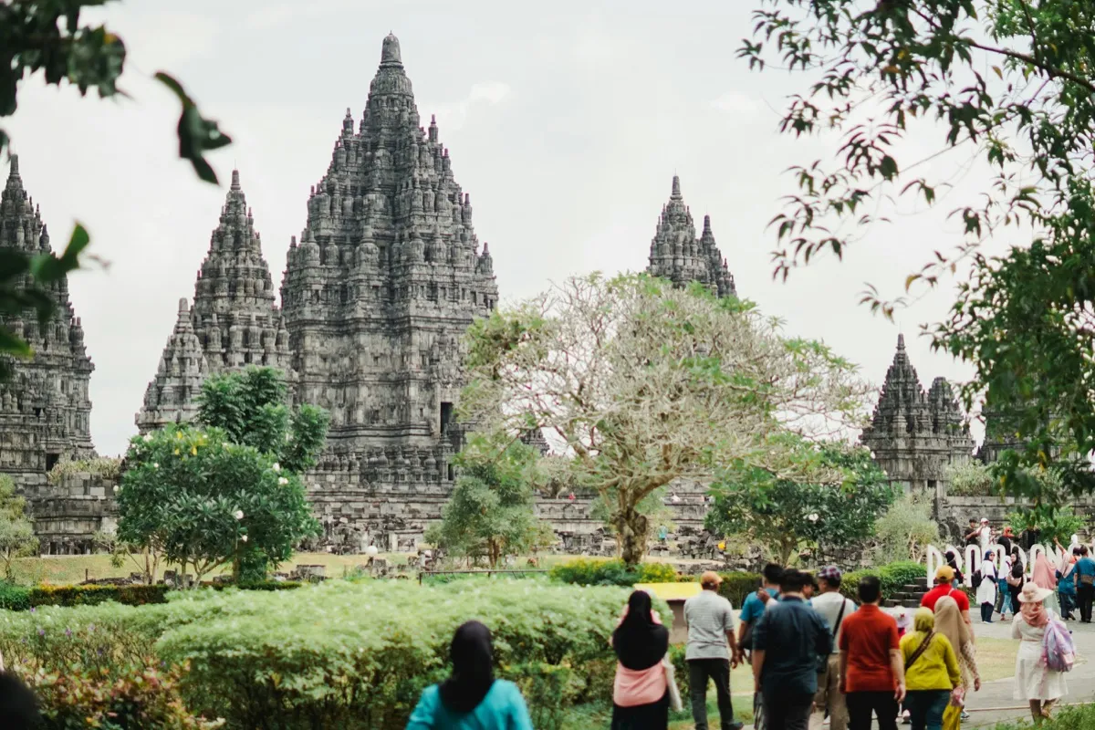 Visitors in modest tropical clothing walk through Prambanan's central courtyard — Java's active Hindu religious site requires long trousers or skirts, sunhats, and closed shoes